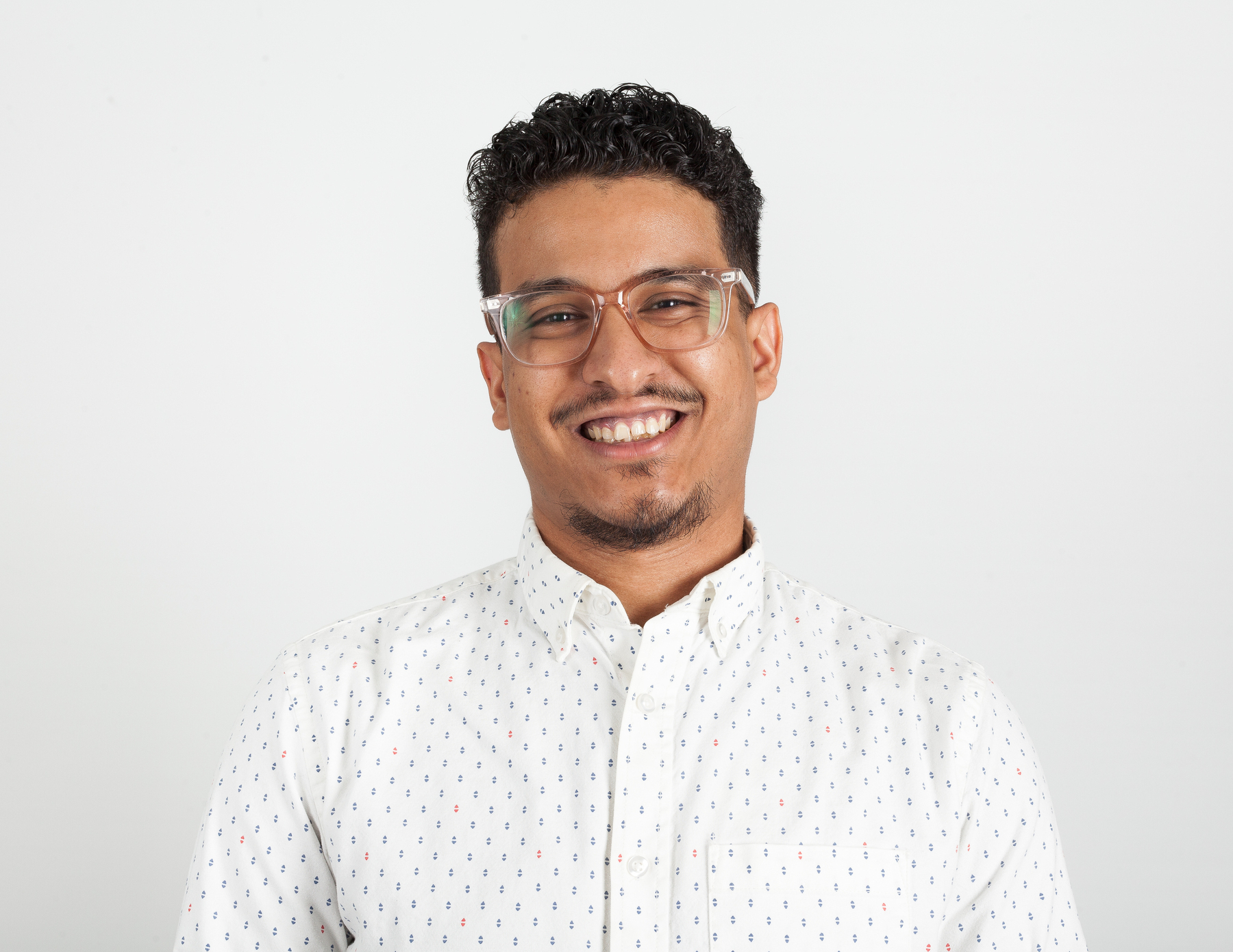 Auto-generated description: A person wearing glasses and a patterned white shirt is smiling against a plain background.